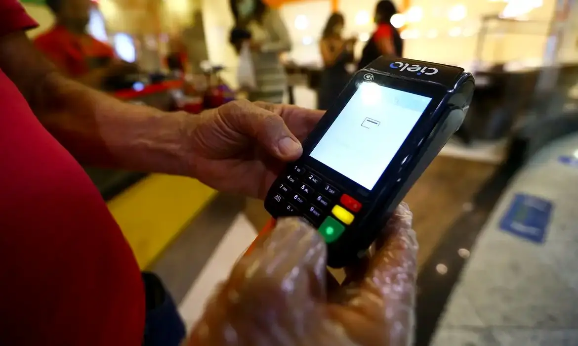 Pessoa realizando pagamento com cartão no terminal de ponto de venda (POS) em ambiente de loja ou restaurante, foco na máquina de cartão e na mão do cliente.
