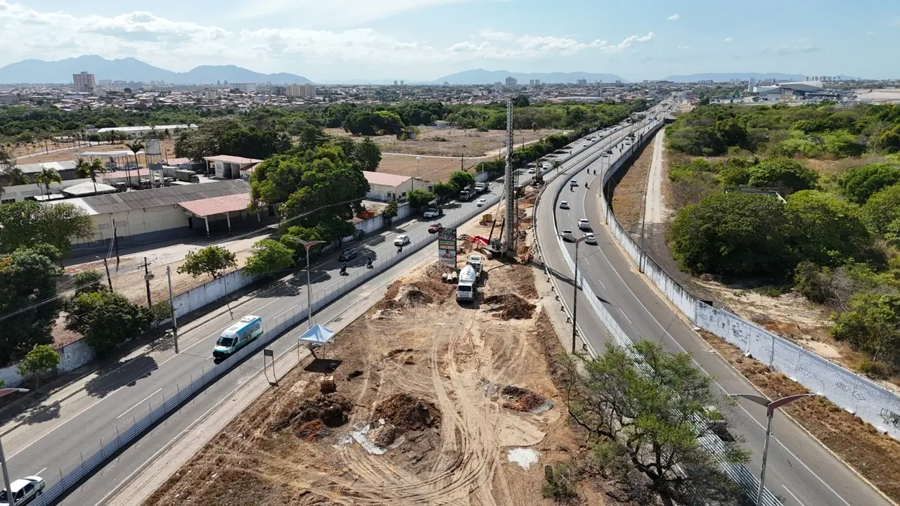 Vista aérea de uma avenida em Fortaleza mostrando obras de construção no canteiro central. Há máquinas e caminhões trabalhando, enquanto carros seguem pelas duas vias paralelas. À esquerda, há prédios baixos e áreas arborizadas; à direita, uma pista expressa cercada por vegetação. Ao fundo, vê-se a cidade com prédios e montanhas no horizonte.