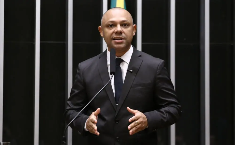 Homem pardo careca com terno preto discursando em palanque com microfone.