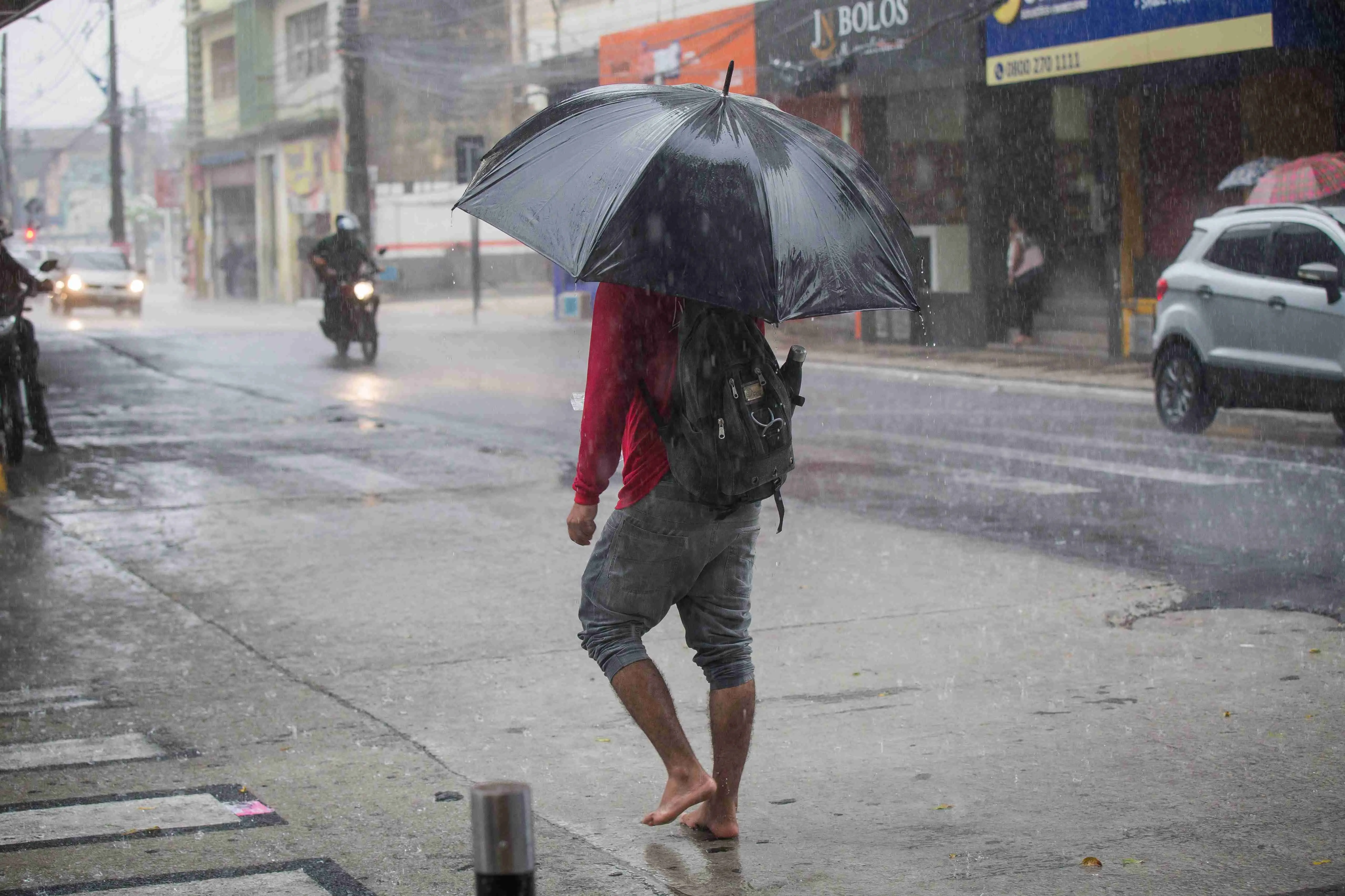 Pessoa descalça com guarda-chuva preto caminhando em rua urbana durante chuva forte.