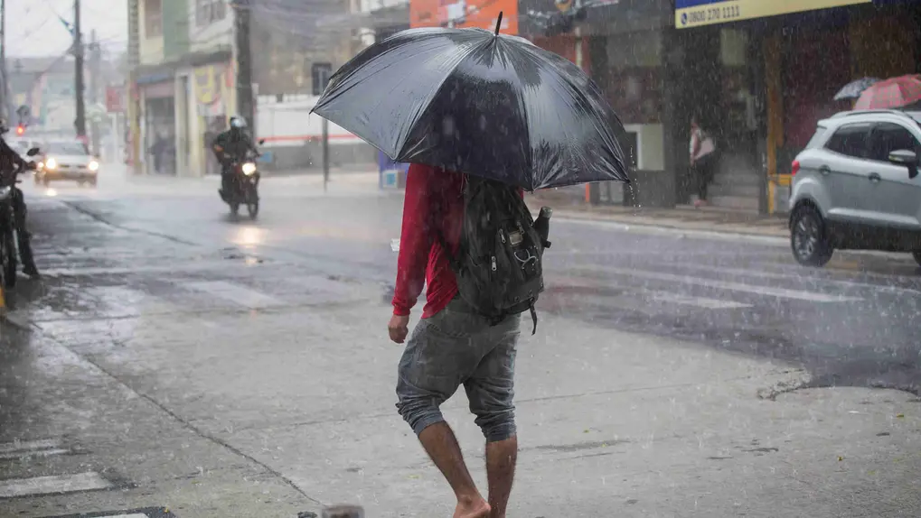 Pessoa descalça com guarda-chuva preto caminhando em rua urbana durante chuva forte.