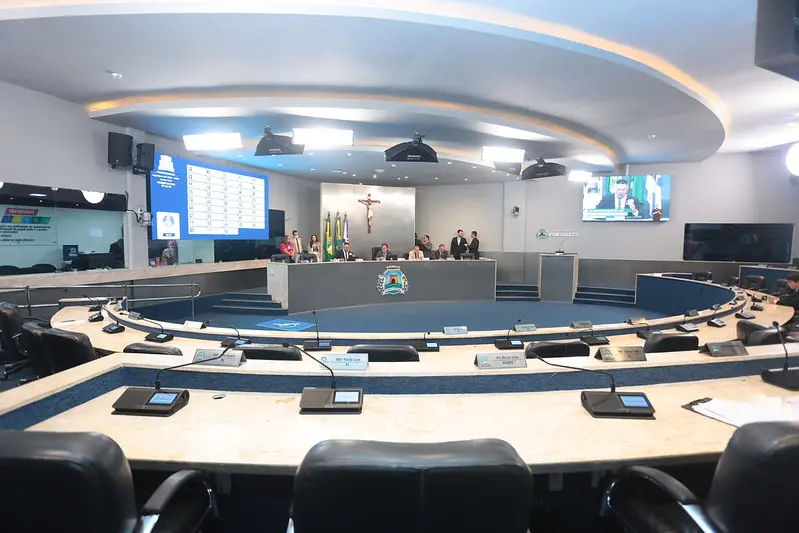 Plenário da Câmara Municipal de Fortaleza visto de forma ampla, com mesas em formato semicircular, microfones individuais, painel eletrônico de votação ao fundo, bandeiras oficiais e crucifixo na parede, durante sessão legislativa.