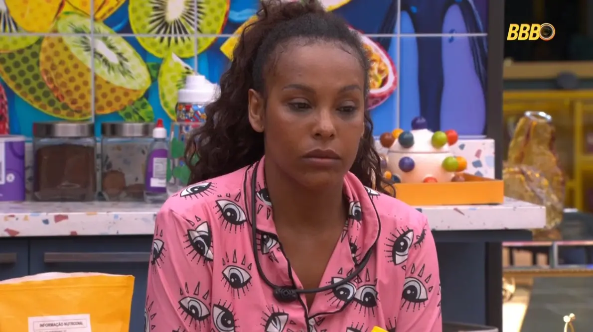 Participante do reality show BBB Brasil com expressão pensativa na cozinha decorada colorida