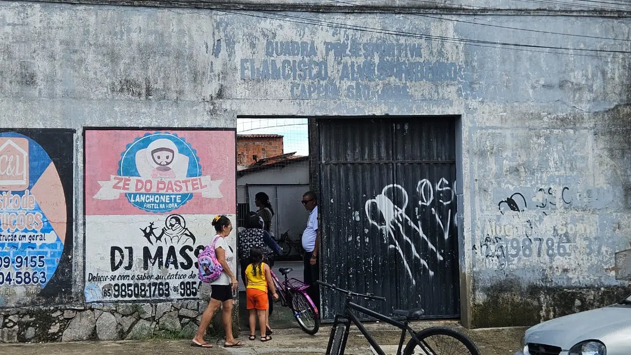 Fachada de um edifício com pintura descascada, exibindo múltiplos cartazes e grafites, com pessoas interagindo perto de uma porta aberta e uma bicicleta estacionada.