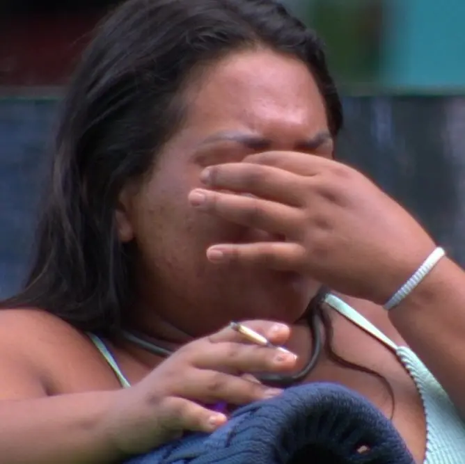 Pessoa sentada ao ar livre segurando um cigarro com uma das mãos, usando uma blusa de alças claras e uma pulseira fina no pulso. O fundo mostra tons desfocados de verde e azul.