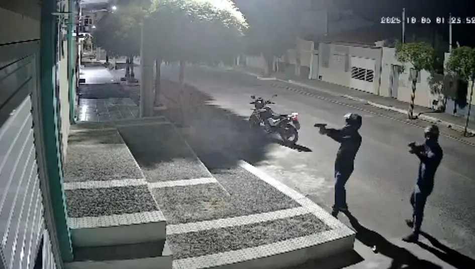 Imagem de câmera de segurança noturna mostrando dois homens armados com pistolas, um deles com capacete, em uma rua escura com uma motocicleta estacionada ao fundo.