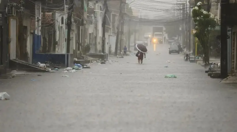 Uma rua urbana completamente alagada durante uma forte chuva, com água cobrindo toda a via; uma pessoa caminha ao centro segurando um guarda-chuva, há lixo espalhado nas laterais, prédios residenciais dos dois lados e um veículo com faróis acesos ao fundo sob céu cinzento.