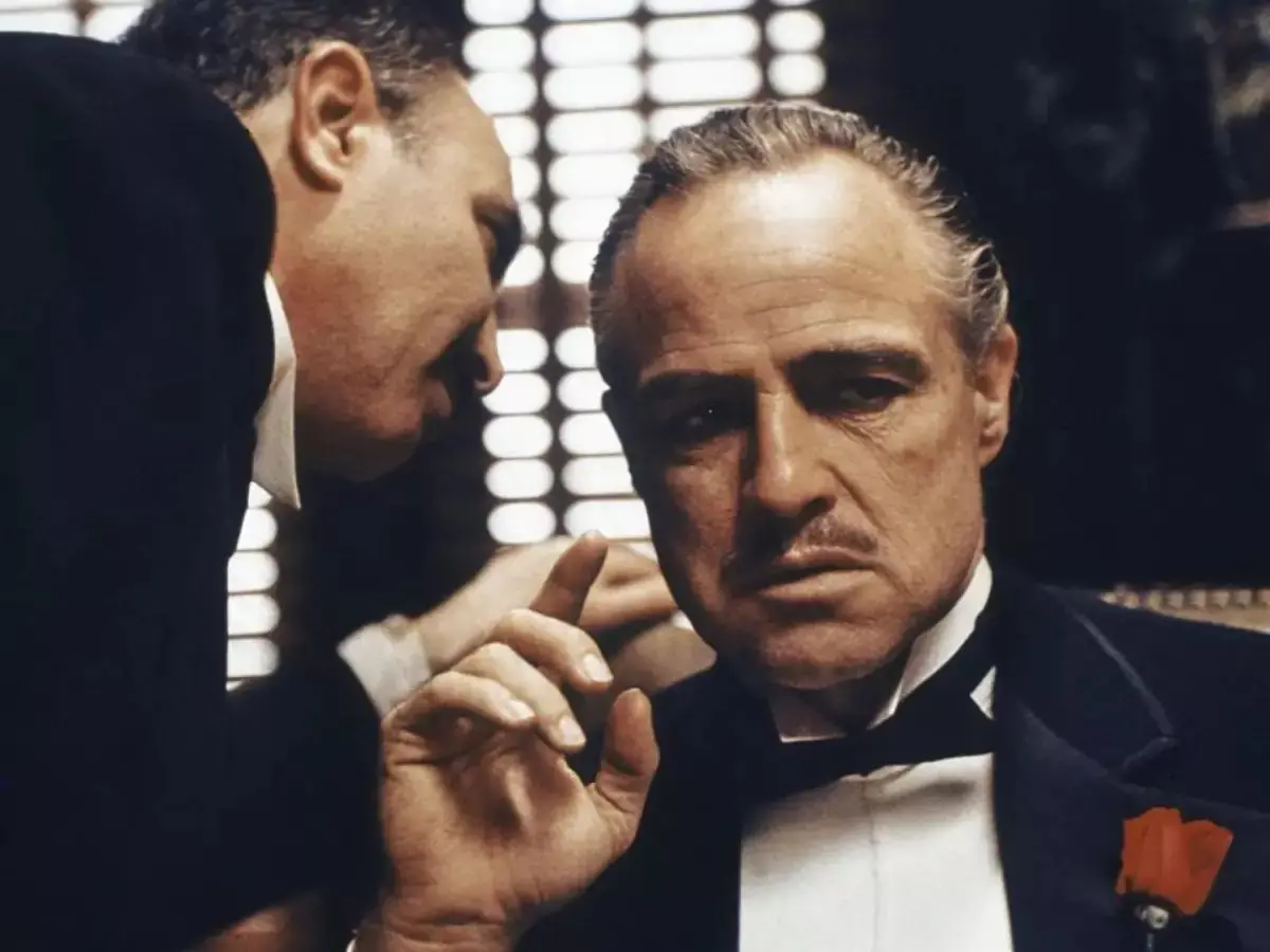 Cena de filme clássico com atores em traje formal, com expressão séria, destacando-se o personagem principal em um ambiente com luzes brilhantes ao fundo.