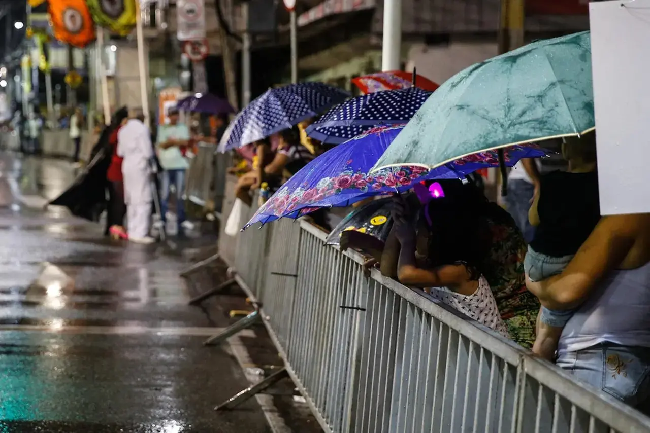 Pessoas assistindo a um evento noturno na rua sob chuva, segurando guarda-chuvas coloridos atrás de barreiras metálicas.