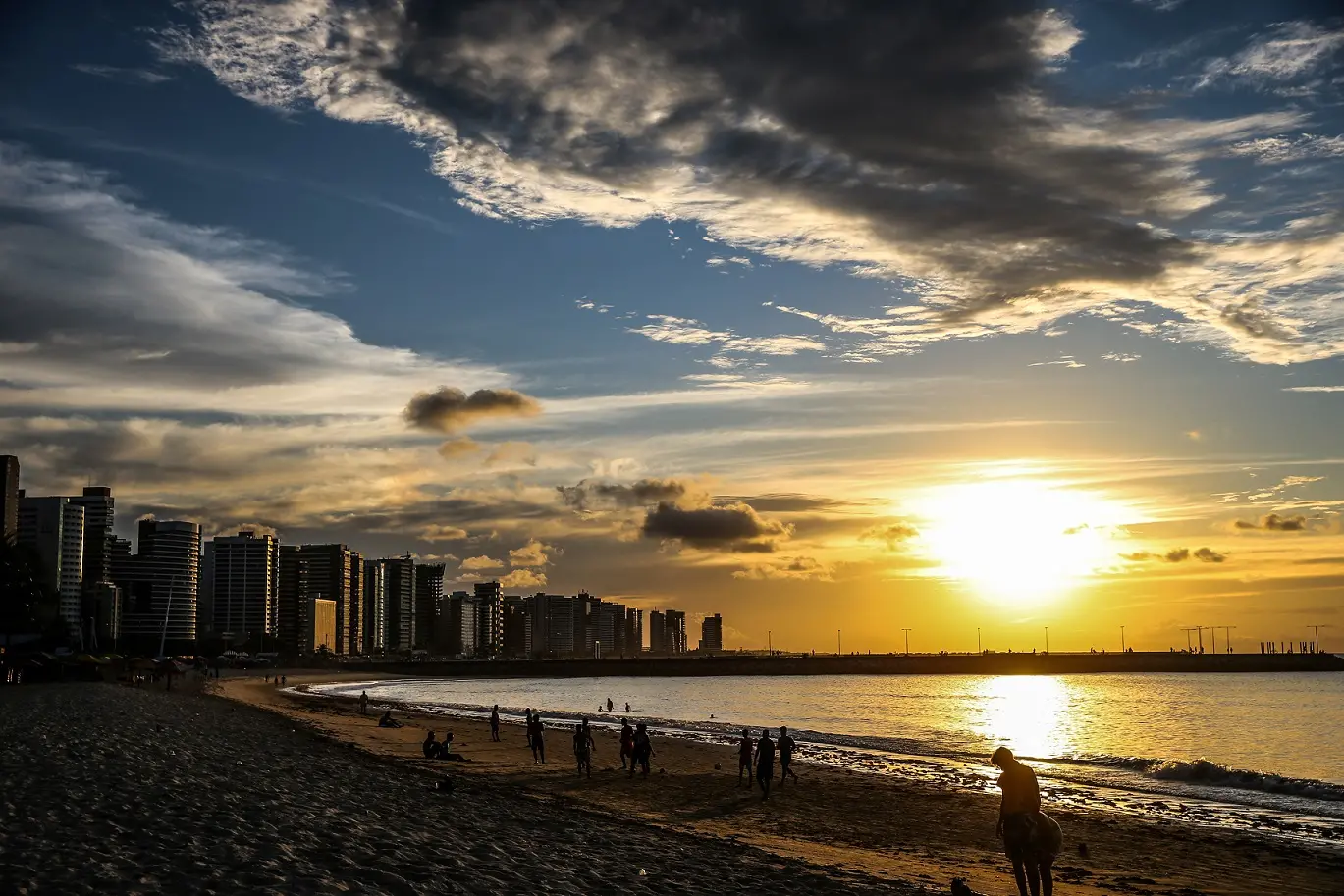 Vista panorâmica da Praia de Iracema, em Fortaleza, ao entardecer, com o sol brilhante refletindo na água, céu com nuvens e a silhueta da cidade costeira.