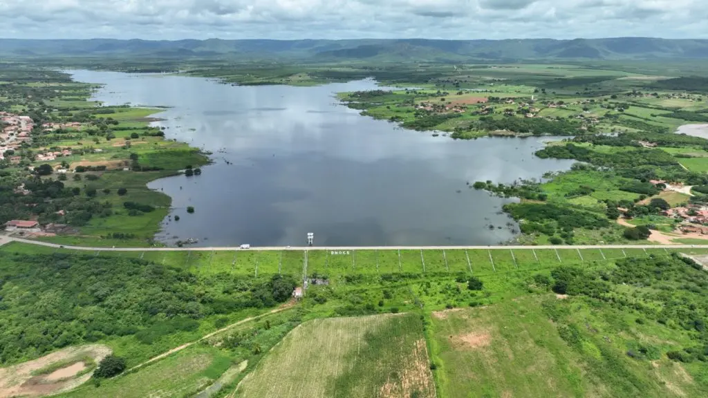 Vista aérea de uma grande barragem, o açude Realejo, com a inscrição DNOCS em seu muro, cercada por campos verdes, vegetação densa, pequenas casas e montanhas distantes sob um céu nublado.