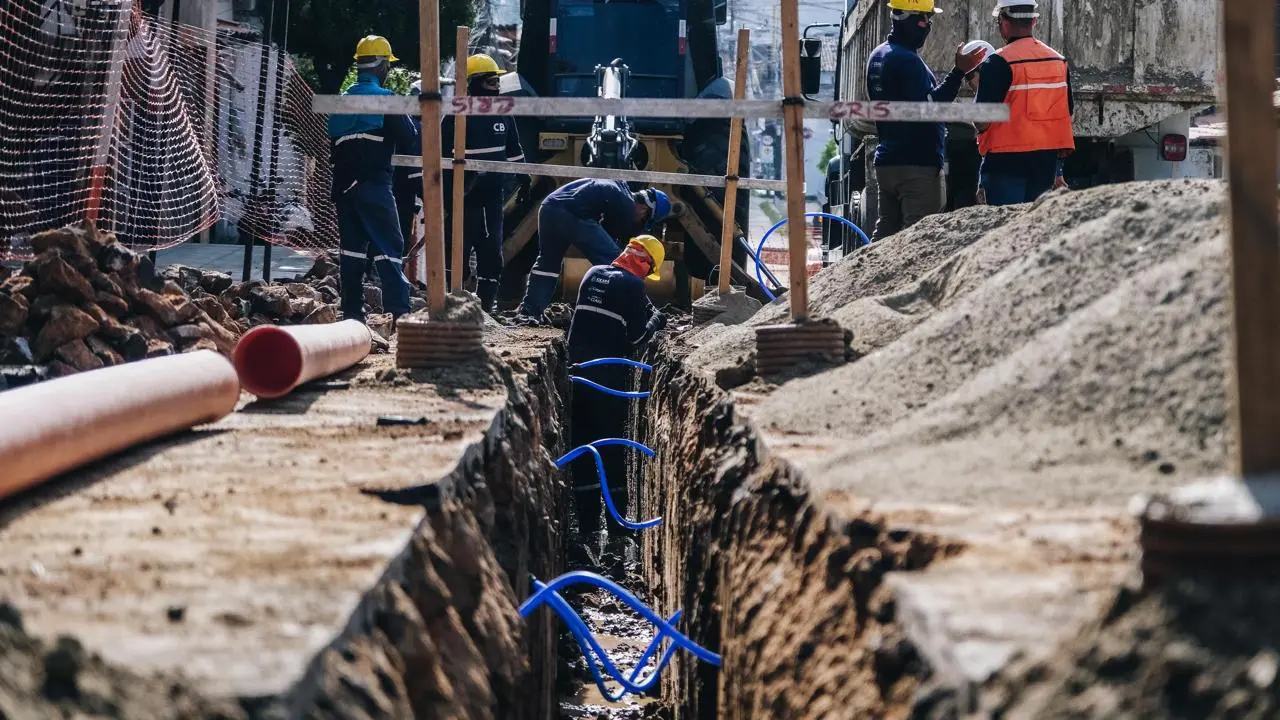 Homens trabalham em obra de vala aberta em chão para colocação de canos de esgotamento sanitário.