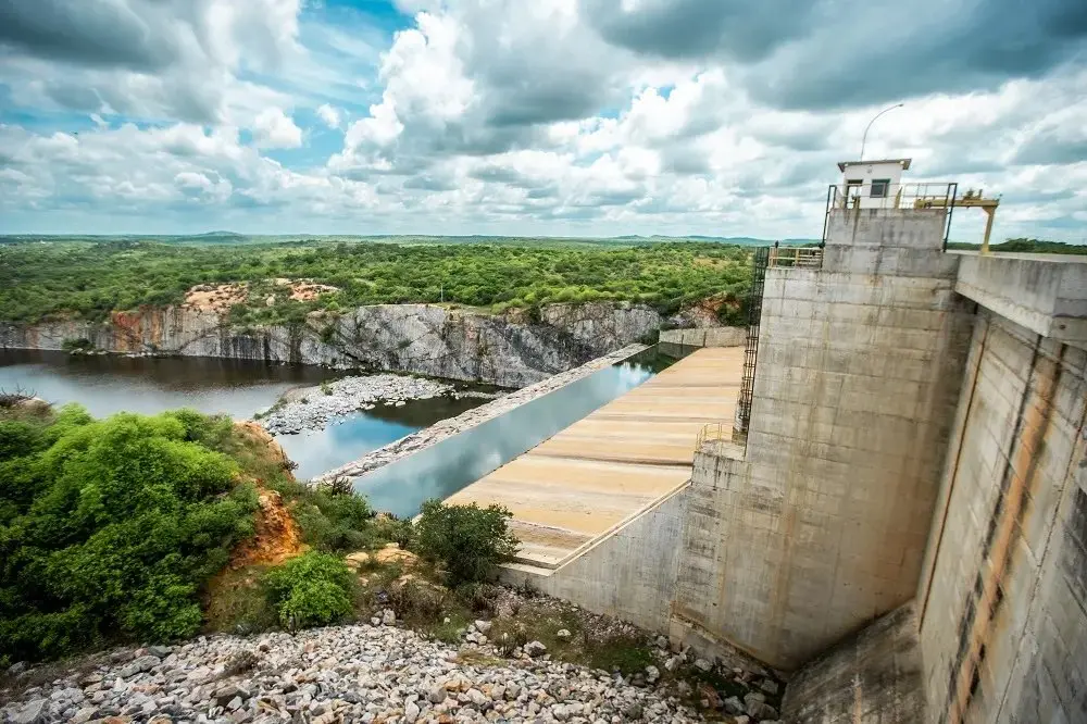 Uma grande barragem de concreto se estende ao lado de um reservatório de água, com um canal que atravessa uma paisagem rochosa e arborizada sob um céu parcialmente nublado.