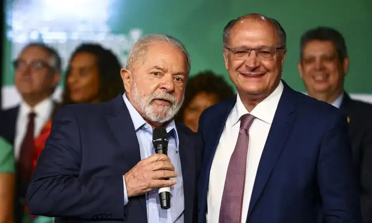 Fotografia em plano médio do presidente Luiz Inácio Lula da Silva e do vice-presidente Geraldo Alckmin, lado a lado, sorrindo. Lula, à esquerda, segura um microfone e veste um terno azul-marinho com camisa azul clara. Alckmin, à direita, veste terno azul-marinho, camisa branca e gravata lilás. Ao fundo, outras pessoas aparecem desfocadas em um ambiente com painéis verdes.