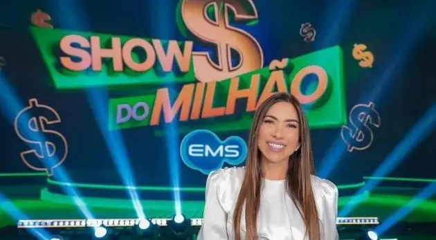 Patrícia Abravanel é uma mulher branca de cabelo castanho claro, longo e liso. Na foto, ela sorri em frente à logo do Show do Milhão.