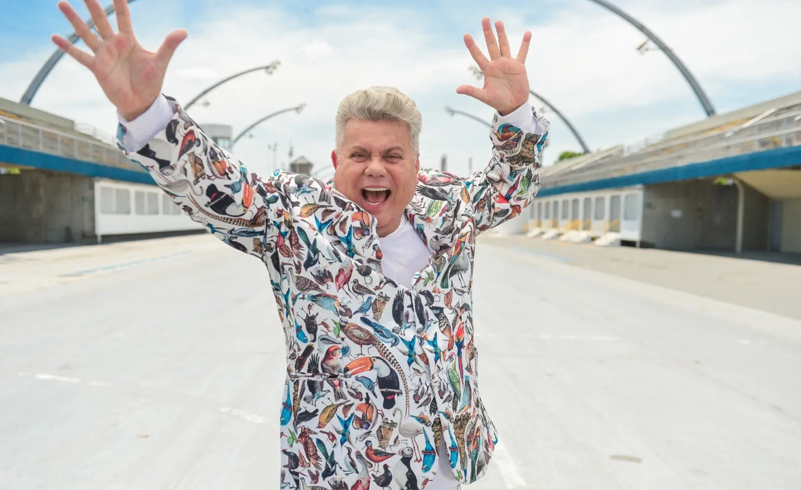 Milton Cunha é um homem idoso de cabelo branco e na foto está de braços abertos no sambódromo.