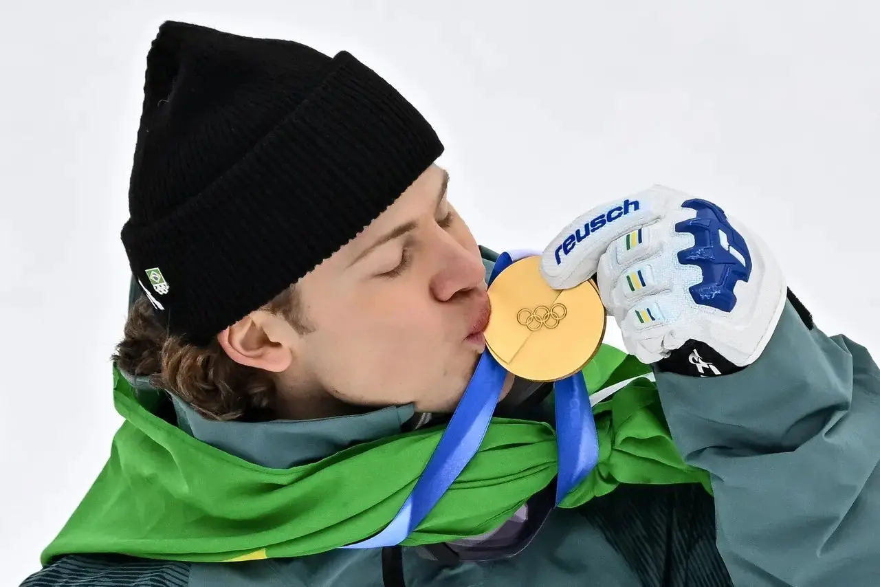 Lucas Pinheiro está vestindo roupas de inverno e bandeira verde e amarela segura uma medalha de ouro olímpica e a leva à boca, usando luvas esportivas brancas e um gorro preto. Imagem usada em matéria onde Lula parabeniza atleta pela conquista.