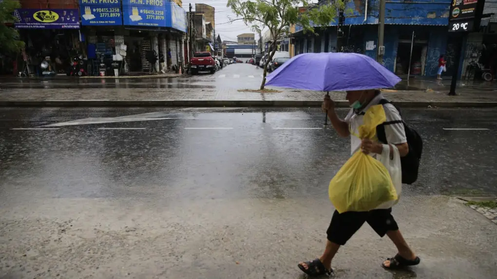 Pessoa atravessando a rua com guarda-chuva na chuva, carregando sacolas de compras, em um dia chuvoso na cidade, com trânsito e lojas ao fundo.