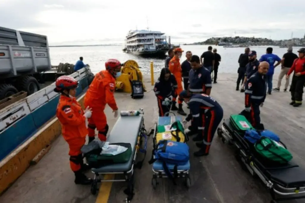 Equipes de resgate com uniformes laranja e azul organizam macas e equipamentos médicos em cais às margens de rio, enquanto embarcação permanece ao fundo, durante operação de atendimento a ocorrência.