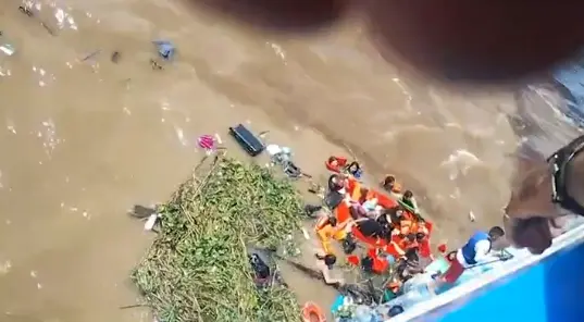 Vista aérea de equipe de resgate com coletes salva-vidas laranja prestando socorro a pessoas em área de rio com água barrenta, próximas a destroços e vegetação flutuante, ao lado de embarcação.