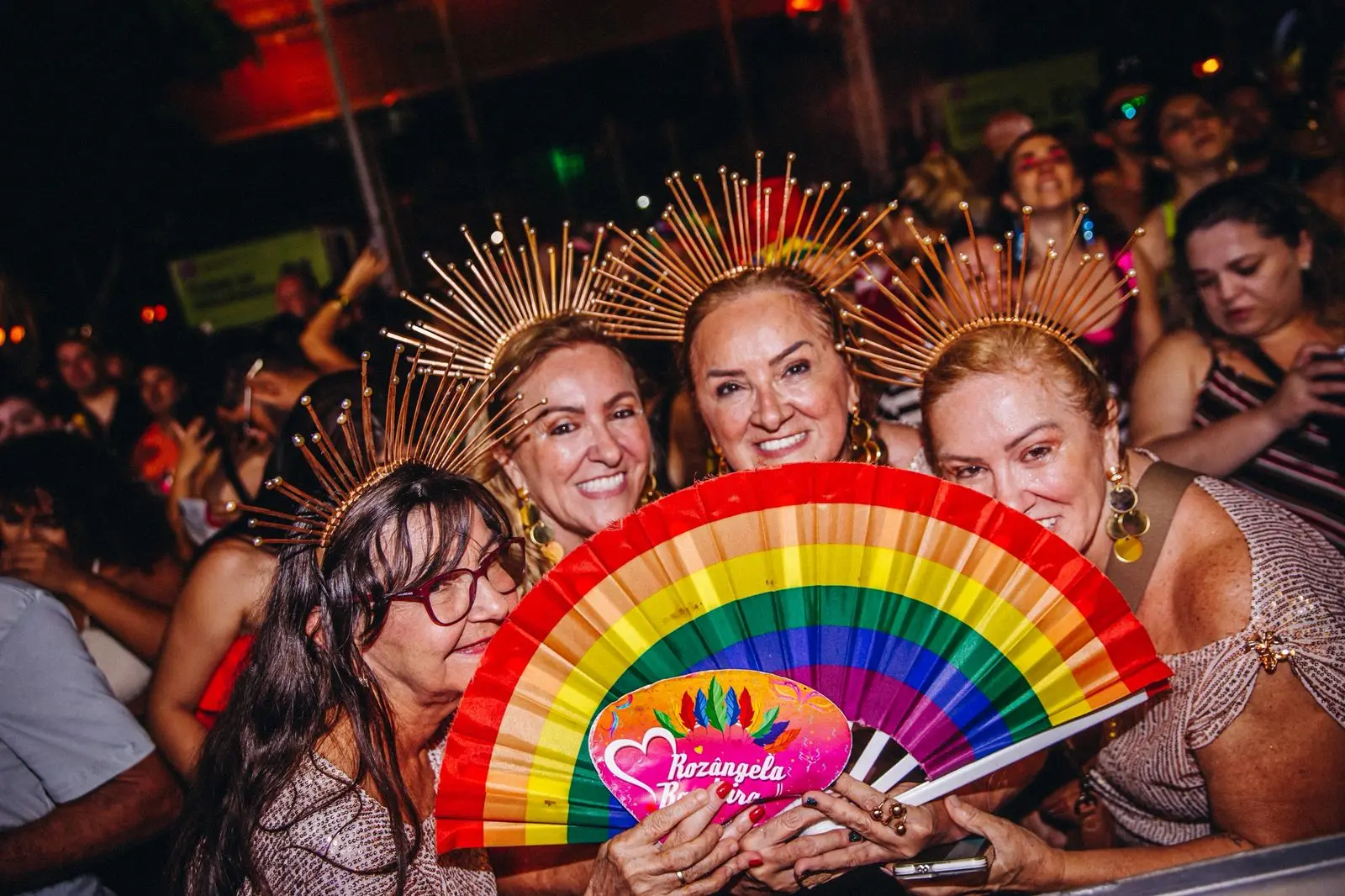 Quatro mulheres posam sorrindo em meio à multidão; usam adereços dourados na cabeça e seguram leque com as cores do arco-íris durante evento noturno.