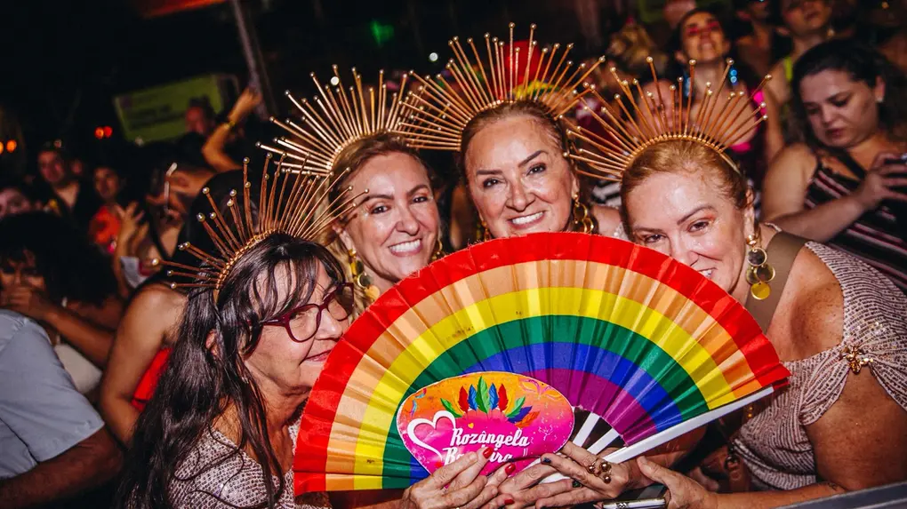Quatro mulheres posam sorrindo em meio à multidão; usam adereços dourados na cabeça e seguram leque com as cores do arco-íris durante evento noturno.
