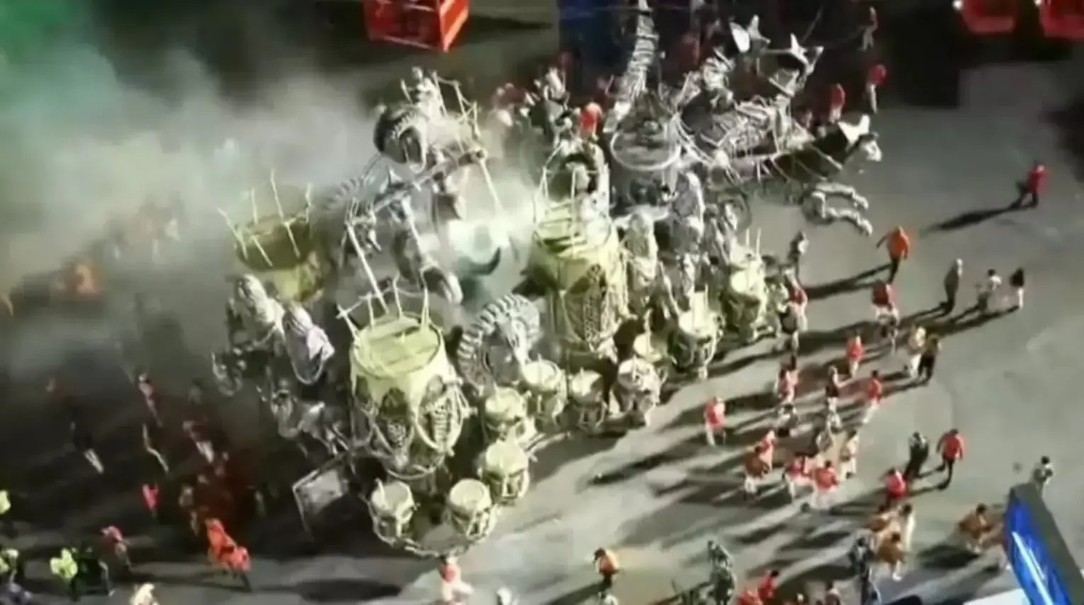 Um dos carros alegóricos da escola em meio à fumaça. A imagem é aérea e é possível ver alguns integrantes da escola no entorno do carro.