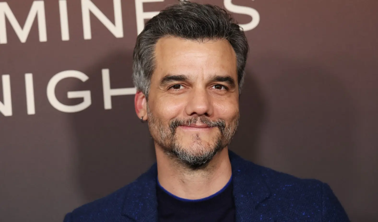 Wagner Moura é um homem branco de meia idade de cabelo preto quase grisalho e barba da mesma cor. Na foto, ele está sorrindo de boca fechada e usa terno azul com camisa preta por baixo.