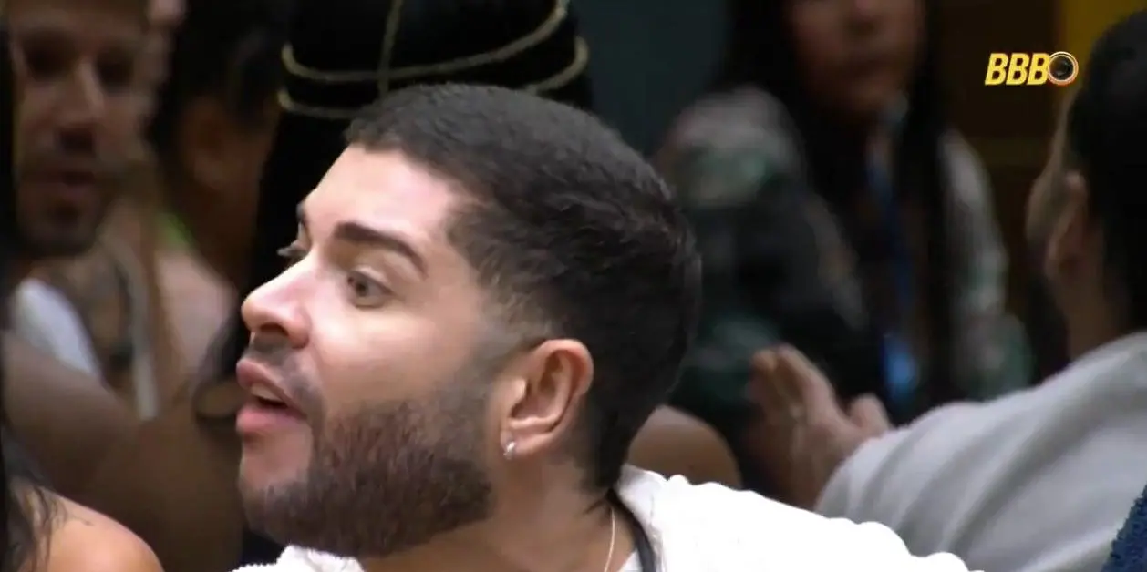 Pessoa sentada em um ambiente interno movimentado, com outras pessoas ao fundo. A iluminação é de estúdio, e há um gráfico colorido na parte inferior da imagem e o logotipo ‘BBB’ no canto superior direito.