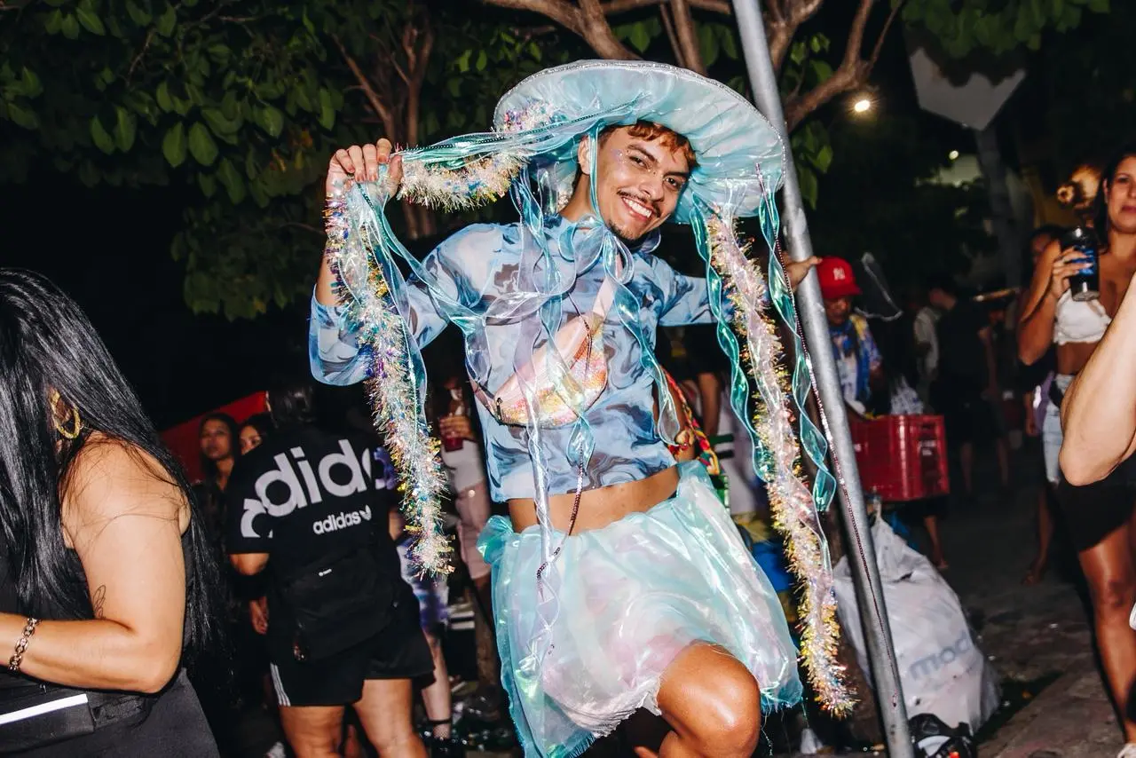 Guilherme fantasiado de água viva. Ele usa chapéu com tentáculos, um top azul estampado e uma saia branca com azul.