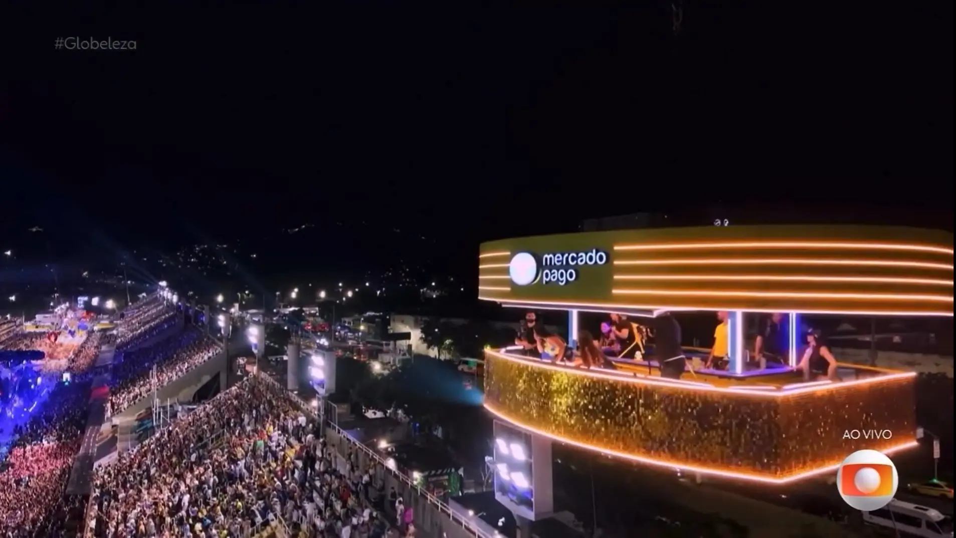Camarote iluminado do BBB com a marca Mercado Pago, suspenso sobre a Sapucaí à noite, com vista para o desfile e uma multidão lotando as arquibancadas; pessoas aparecem no interior do espaço envidraçado, enquanto a transmissão está ao vivo na TV Globo.