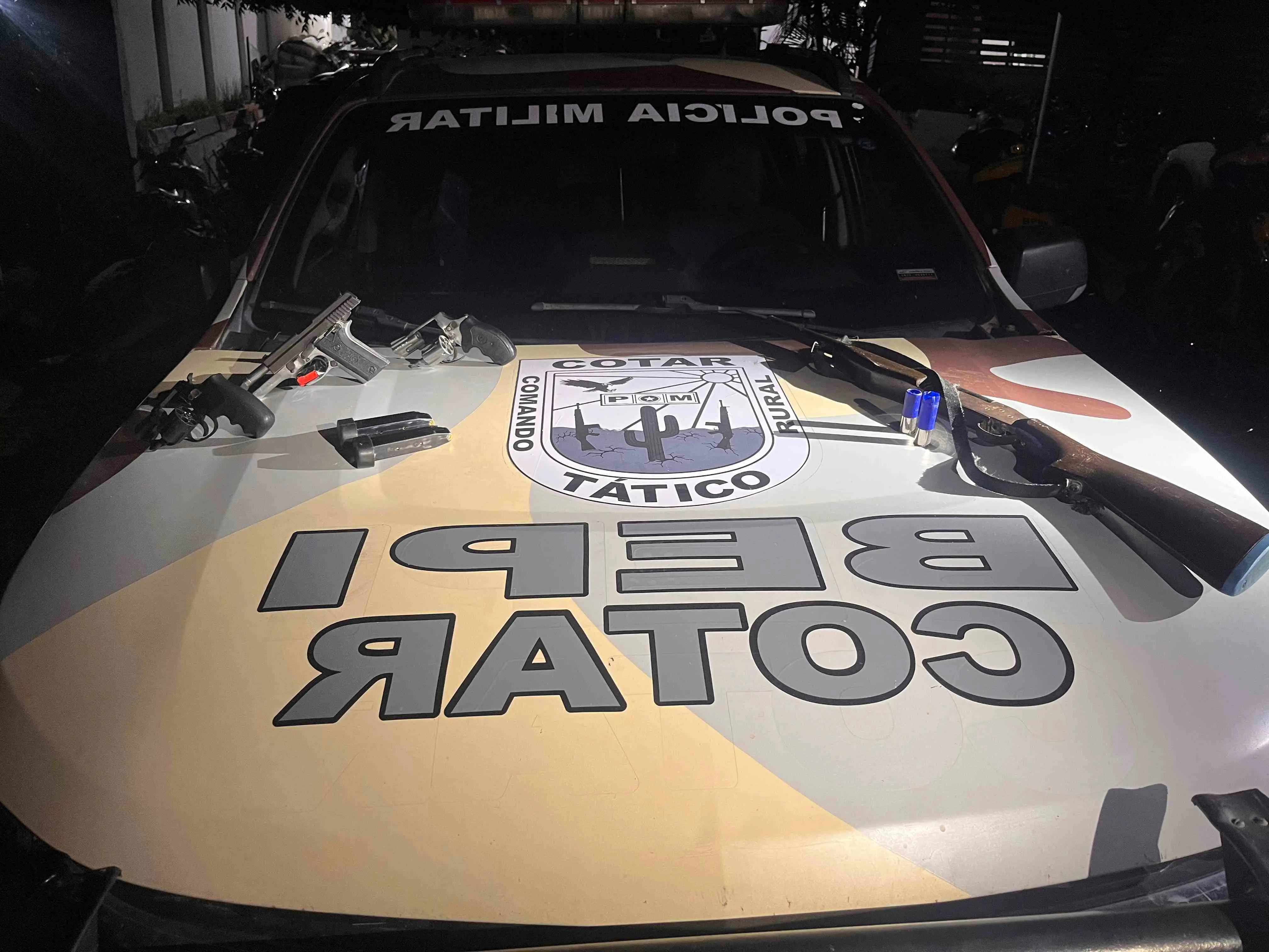 Capô de uma viatura da Polícia Militar do COTAR (Comando Tático Rural) com o brasão e a inscrição “BP COTAR”, exibindo armas apreendidas — dois revólveres, uma pistola, um rifle, carregadores e munições — organizadas sobre o veículo durante a noite.
