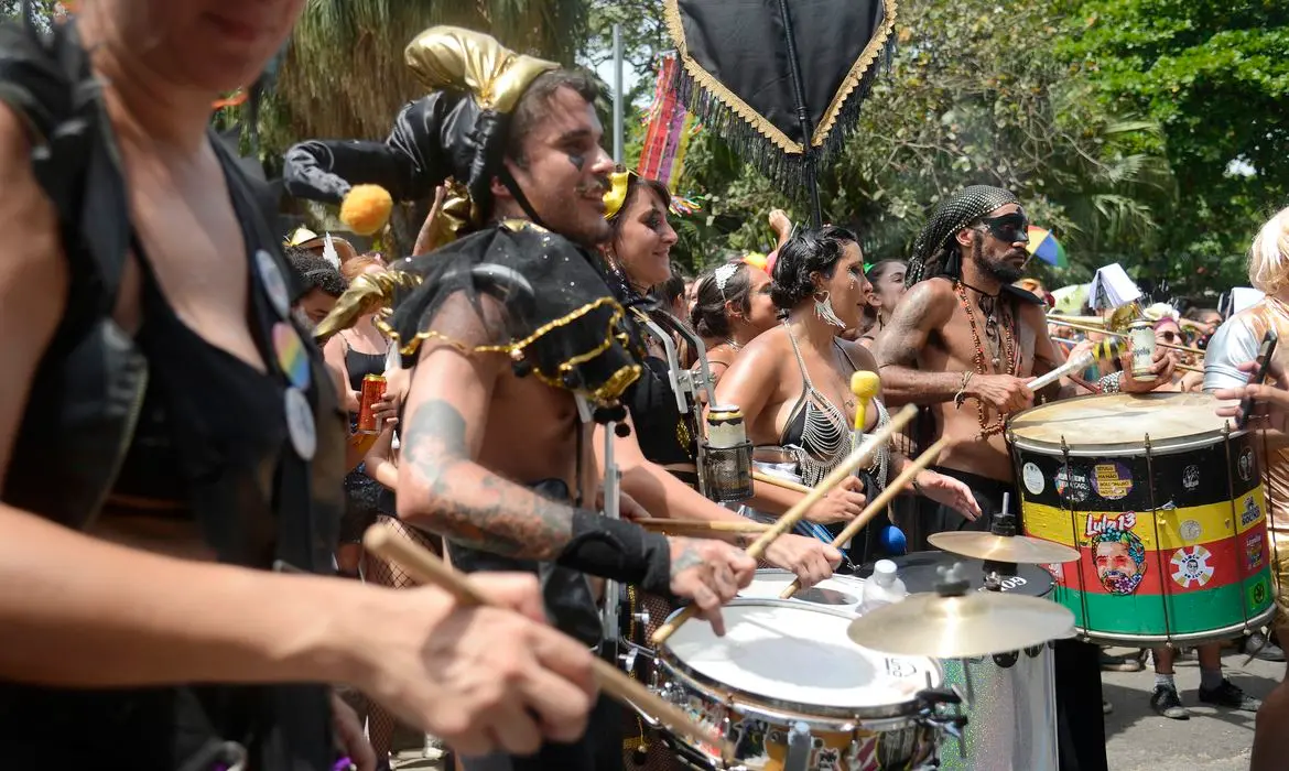 Grupo de percussionistas participando de um desfile ou celebração ao ar livre, com pessoas vestidas com fantasias coloridas e acessórios carnavalescos, cercados por árvores e foliões na rua.