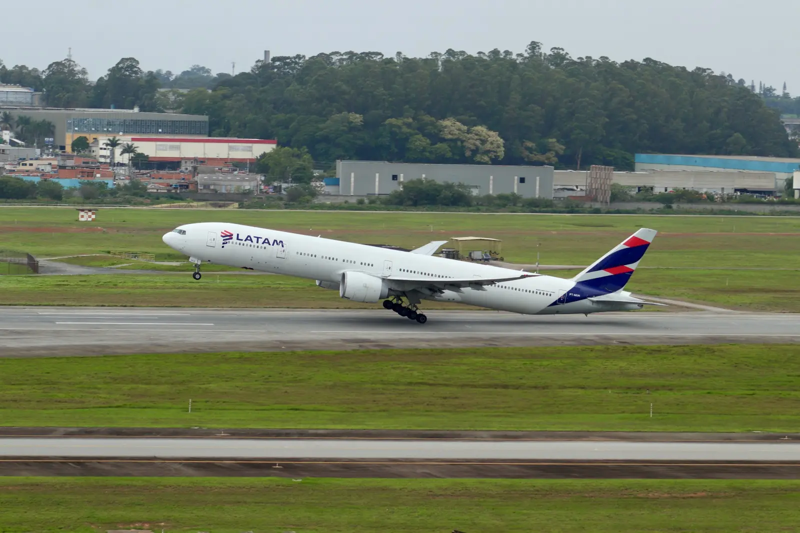 Avião da LATAM acelera na pista durante decolagem no Aeroporto de Guarulhos, com trem de pouso ainda tocando o solo e área urbana ao fundo.