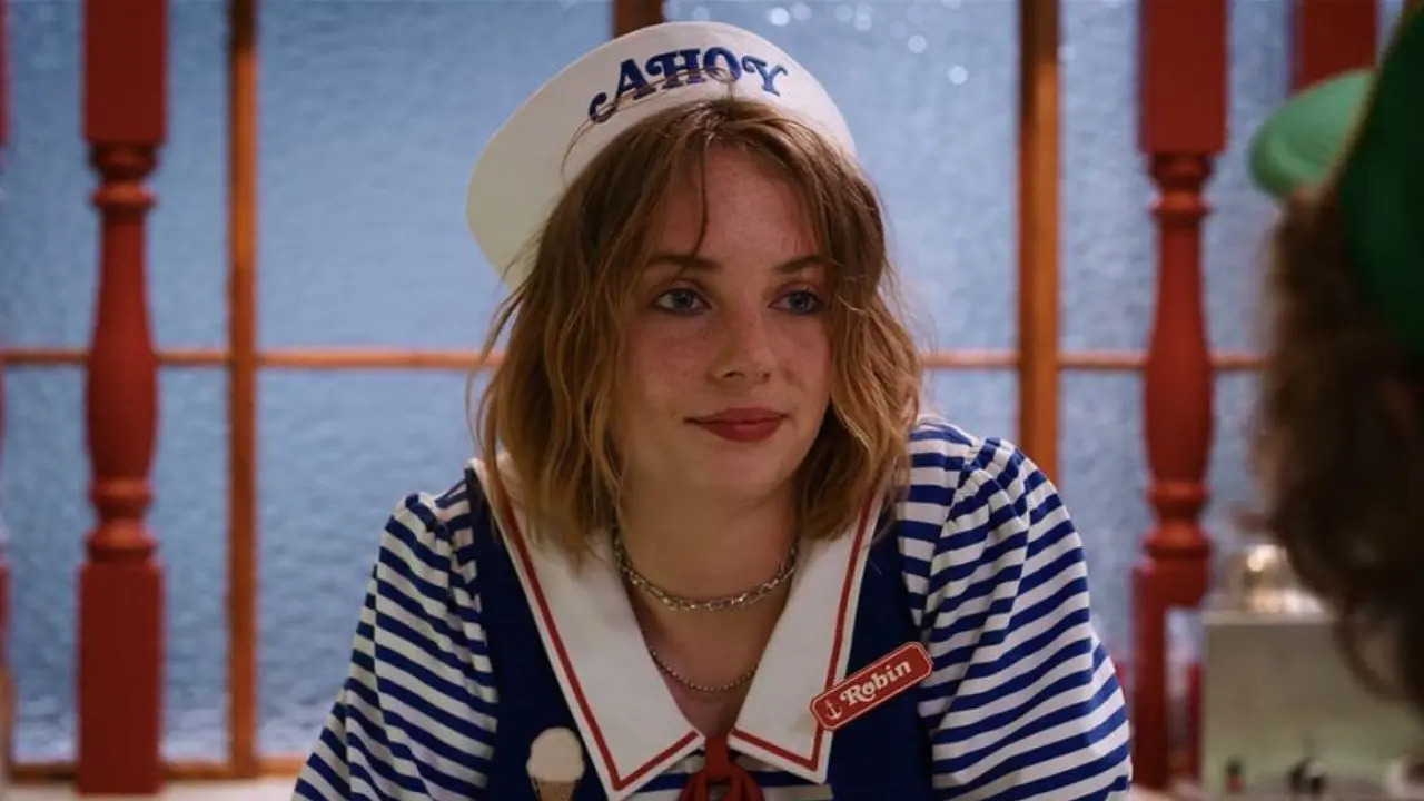 Cena da série 'Stranger Things', com a atriz Maya Hawke sorrindo, usando roupa de marinheira e chapéu de papelão, em cenário colorido.