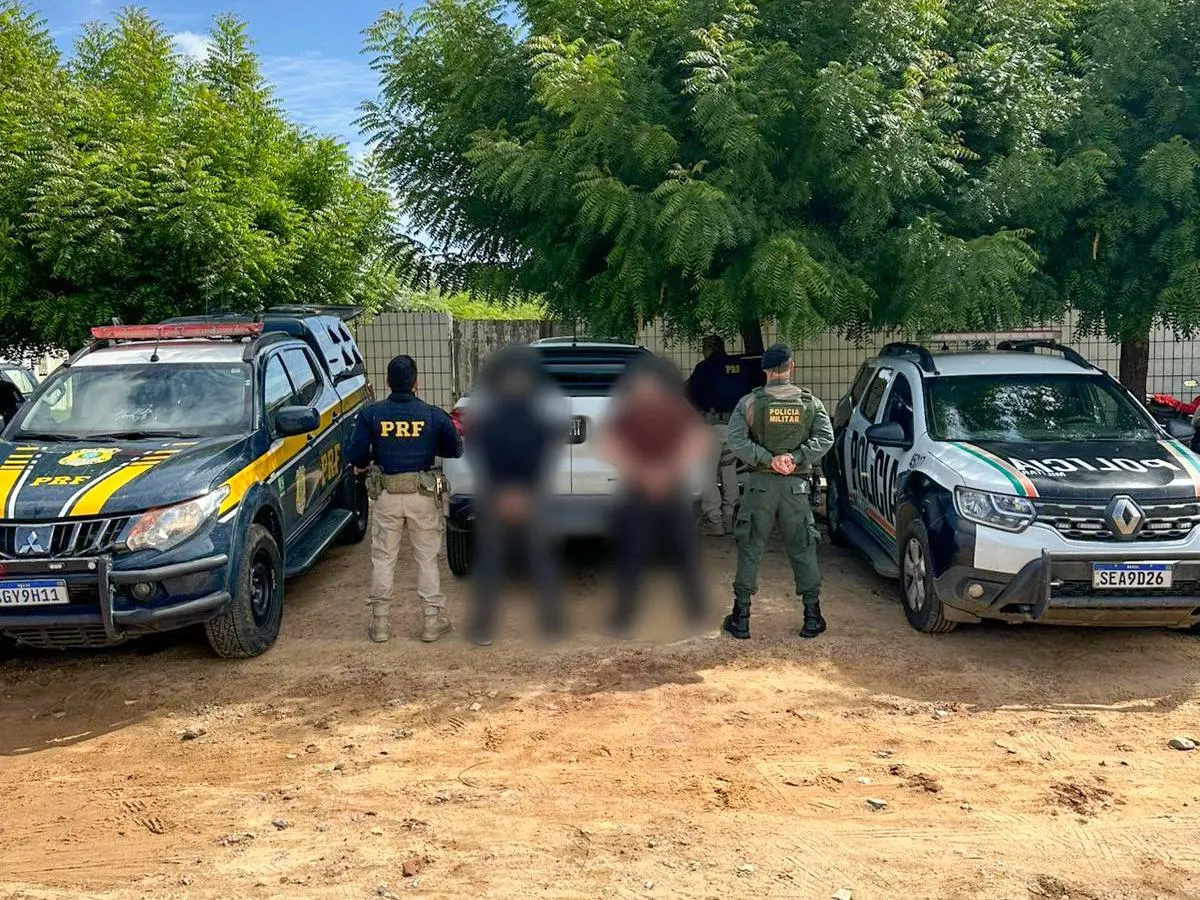 Dois policiais posicionados ao lado de duas pessoas presas, ambas desfocadas, em frente a uma viatura da PRF e uma viatura da Polícia Militar.