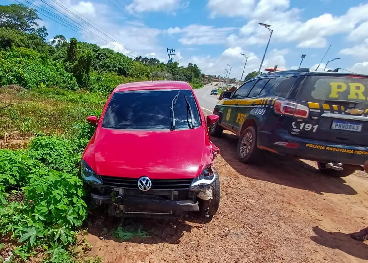 Carro vermelho com a parte dianteira danificada parado no acostamento de uma estrada, ao lado de uma viatura da Polícia Rodoviária Federal. Ao redor há vegetação verde e o céu está parcialmente nublado.