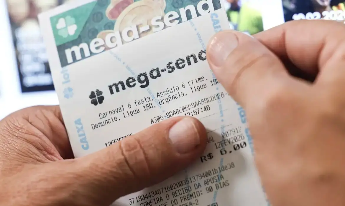 Pessoa segurando um bilhete da Mega-Sena, com o nome da loteria em destaque e o valor da aposta impresso, em frente a uma tela desfocada ao fundo.