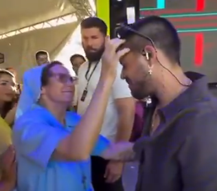 Freira vestida com hábito azul toca a testa de um cantor em cima de um palco, enquanto outras pessoas observam ao redor durante um evento.