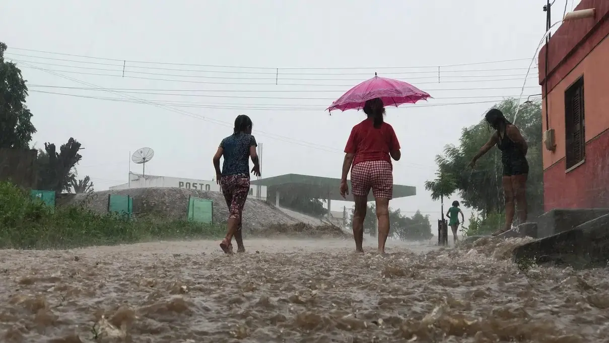 Crianças e adolescente tomam banho de chuva sob o olhar de mulher protegida por um guarda chuva. Em primeiro plano, uma rua de terra coberta por água.