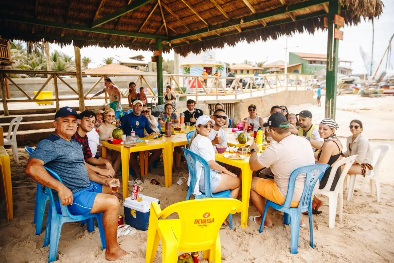 Um grupo de pessoas sentadas sob um toldo improvisado na praia, reunidas ao redor de uma mesa plástica branca com garrafas, copos e lanches. Algumas estão em cadeiras de praia e outras em pé ao fundo. O cenário inclui areia, barcos coloridos ao lado e vegetação baixa próxima às construções ao fundo. O grupo parece relaxado e conversando em um ambiente descontraído à beira-mar.