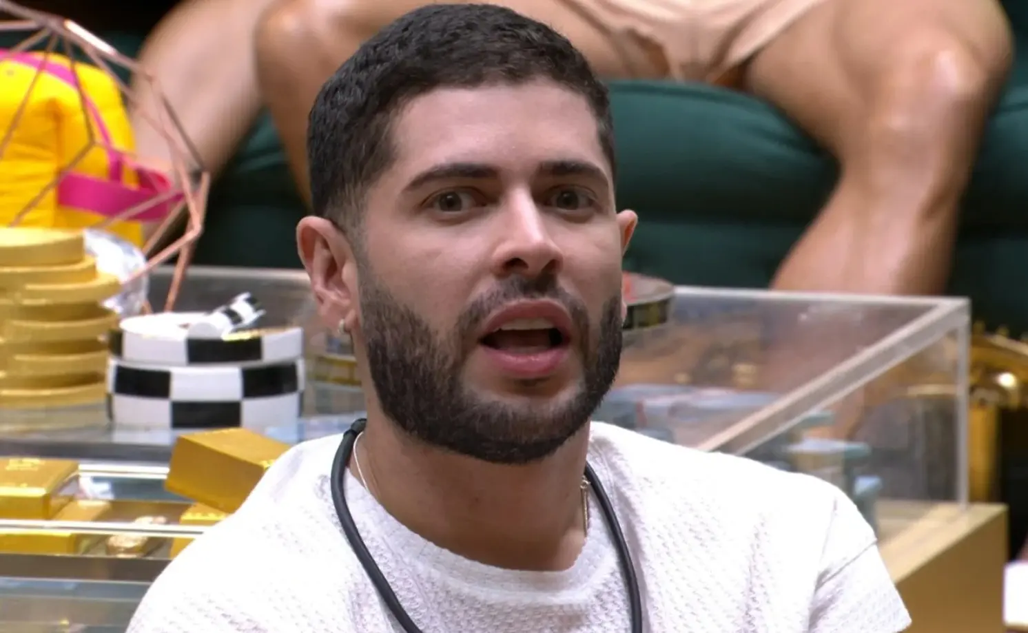 Uma pessoa sentada em um ambiente interno iluminado, vestindo uma camisa de manga longa branca com textura. No fundo, há uma mesa transparente com barras douradas decorativas e algumas fichas de jogo empilhadas. Atrás disso, outra pessoa está sentada em um sofá verde, com as pernas cruzadas. O cenário lembra um espaço de convivência com elementos decorativos brilhantes.