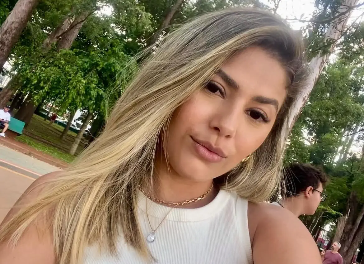 Selfie de Ana Karolina Sousa, uma mulher jovem na faixa de 30 anos com mechas loiras, olhos castanho escuros, sorrindo para a câmera. Ela usa uma camiseta branca e colar de ouro. Ao fundo, há um parque verde.