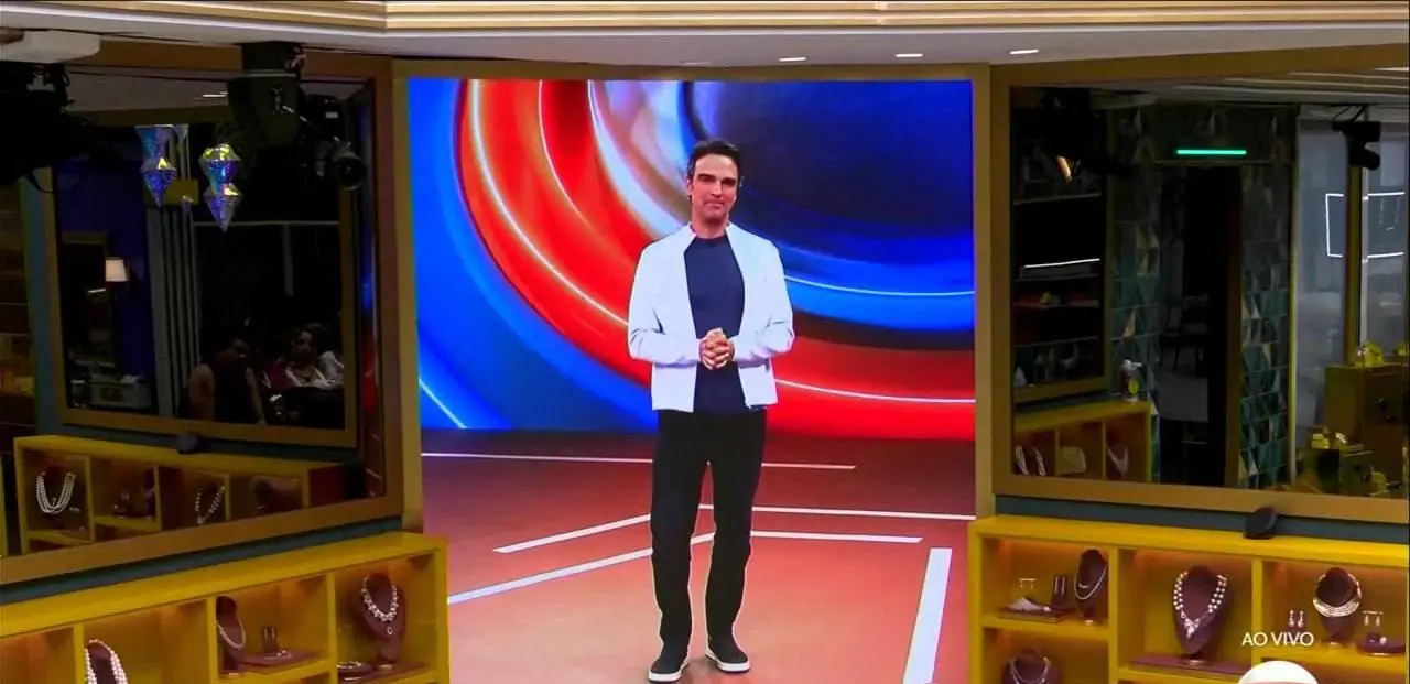 Uma pessoa está em pé no centro de um grande painel eletrônico colorido com formas circulares em tons de azul, vermelho e branco. A pessoa veste uma jaqueta clara, camiseta escura, calça escura e tênis claros, com as mãos unidas à frente do corpo. Ao redor, o ambiente lembra um estúdio iluminado, com vitrines de exibição contendo colares e acessórios nas laterais, além de espelhos refletindo partes do cenário. No canto inferior direito aparece a palavra “AO VIVO”.