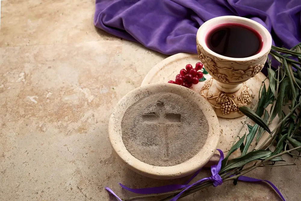 Água benta com cruz em um recipiente de pedra, acompanhado de um cálice decorado com vinho e ramos de oliveira, simbolizando tradições religiosas de fé.