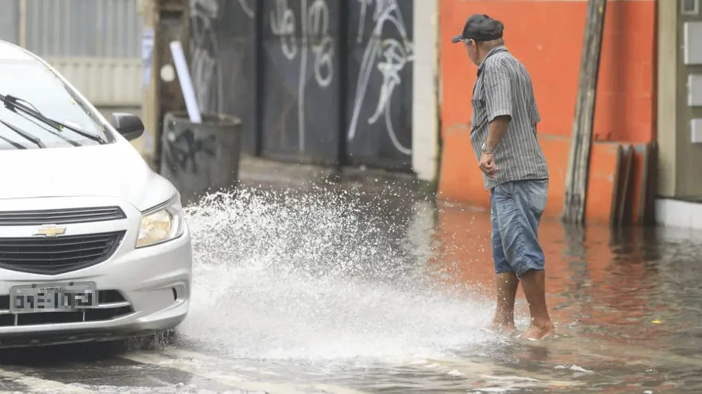 Homem idoso de boné em rua urbana inundada observa carro passar e criar grandes respingos de água.
