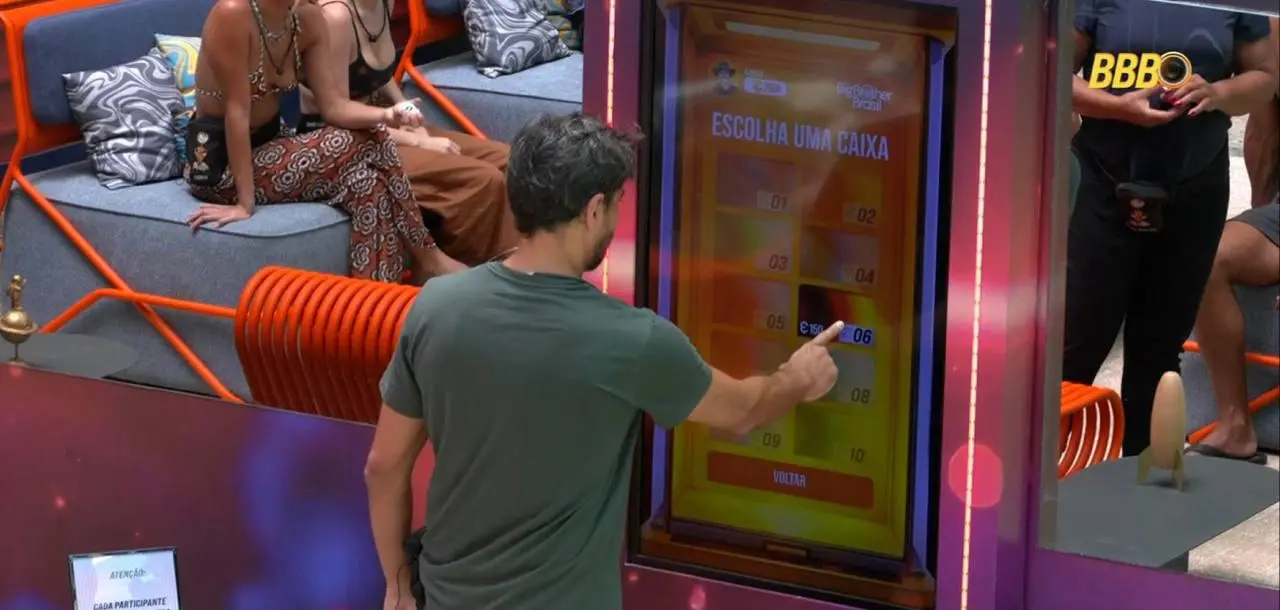 Uma pessoa vista de costas interage com uma tela grande vertical que exibe opções numeradas de 01 a 10 sob o título “Escolha uma caixa”. A pessoa toca na opção 05. Ao fundo, há outras pessoas sentadas em um ambiente colorido com poltronas laranja e cinza. No canto inferior esquerdo há um aviso impresso sobre regras de participação.