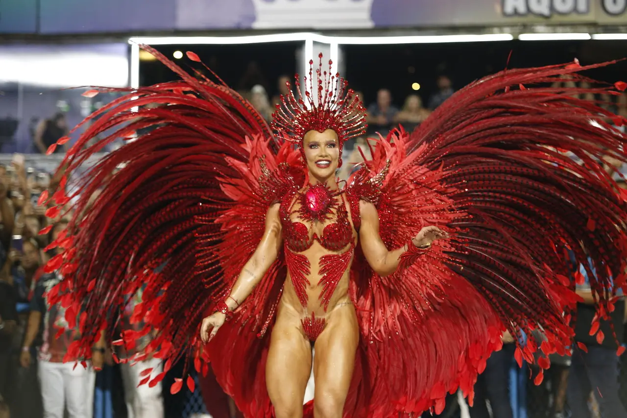 Uma pessoa desfila em uma avenida de Carnaval usando uma fantasia elaborada em tons de vermelho vibrante. A fantasia possui enormes asas compostas por longas penas vermelhas que se abrem amplamente para os lados, além de detalhes brilhantes e ornamentados no corpo. No topo da cabeça, há um adorno alto e estruturado, também vermelho, com elementos que lembram plumas e pedras decorativas. Ao fundo, é possível ver parte do público e da estrutura do sambódromo iluminado.