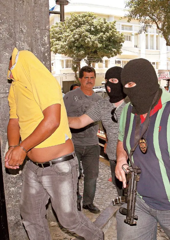 Imagem mostra Evilson Alves Feitosa, de camisa amarela e rosto coberto pela camisa, no momento em que chegava preso na sede da Polícia Civil, no Centro da Capital. O ano era 2012. Ele está escoltado por três policiais, sendo dois de balaclava e um de rosto descoberto, de bigode.