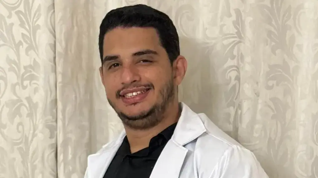 Profissional usando um jaleco branco com o nome “Dr. Yuri Portela – Médico” bordado, posicionado em frente a uma cortina estampada. O jaleco também exibe um brasão no braço com detalhes em dourado. Yuri é o médico que foi preso por assediar aluna em faculdade do Ceará.